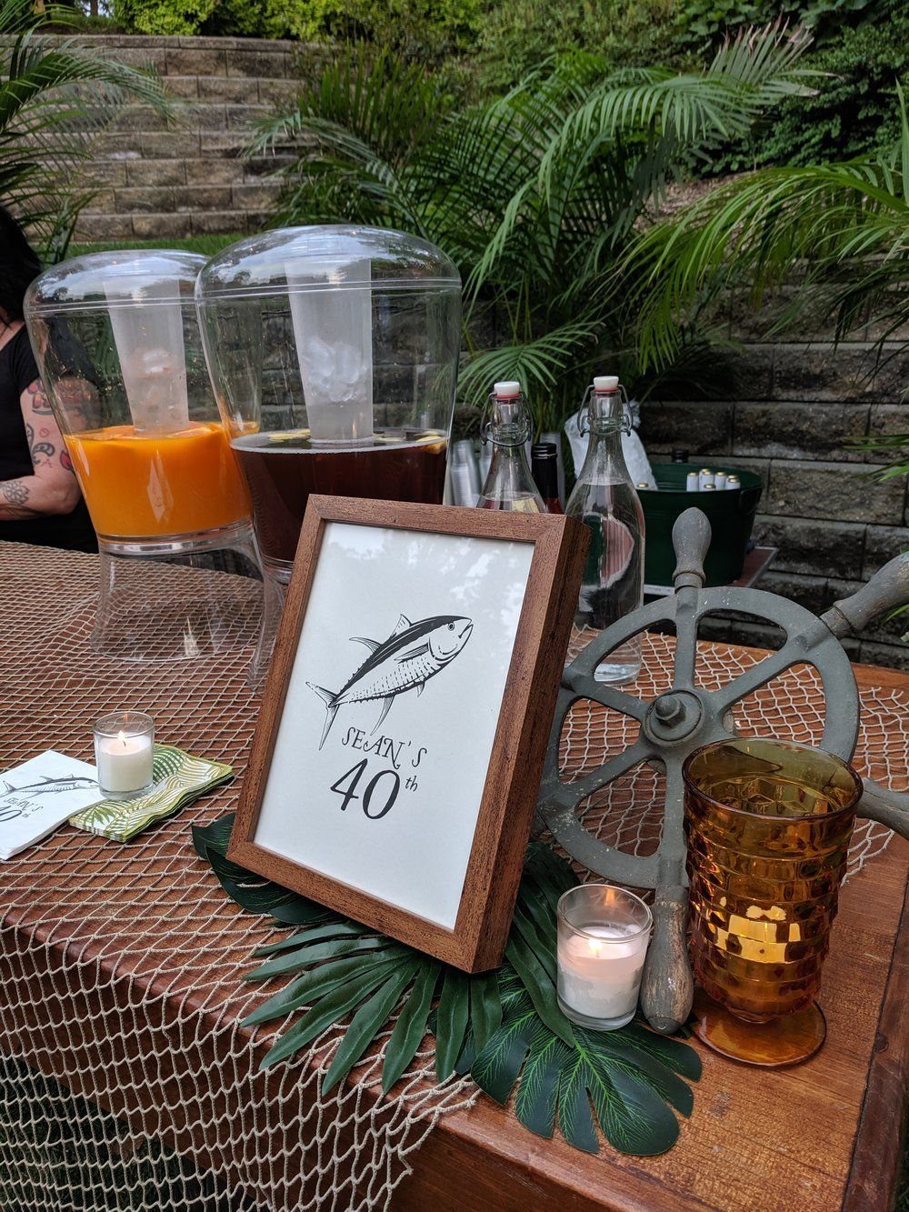 A decorated party table with beverage dispensers, a framed fish sign, and a miniature ship's wheel.