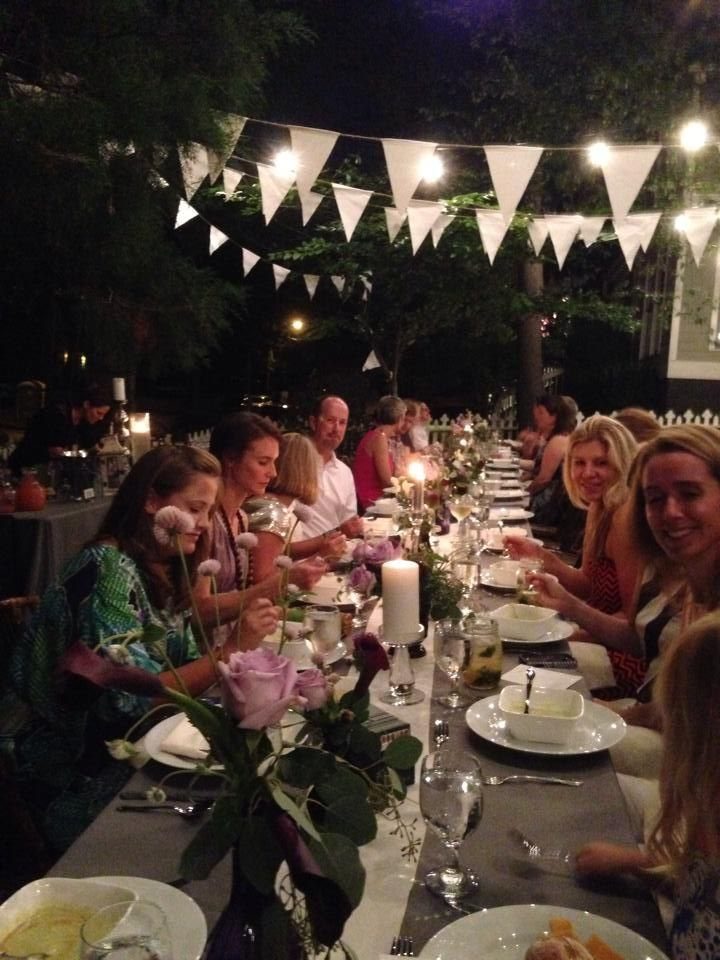 People seated at a long, decorated outdoor dining table with flowers and string lights.