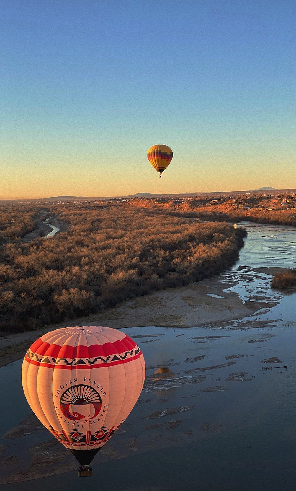Hot air balloons floating over a scenic river valley during a golden sunset.