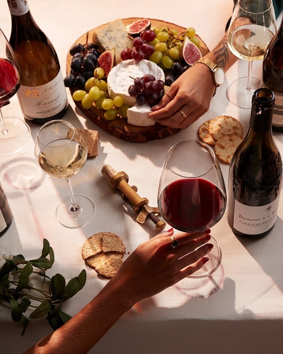 Hands reaching for cheese and grapes on a charcuterie board surrounded by wine glasses.