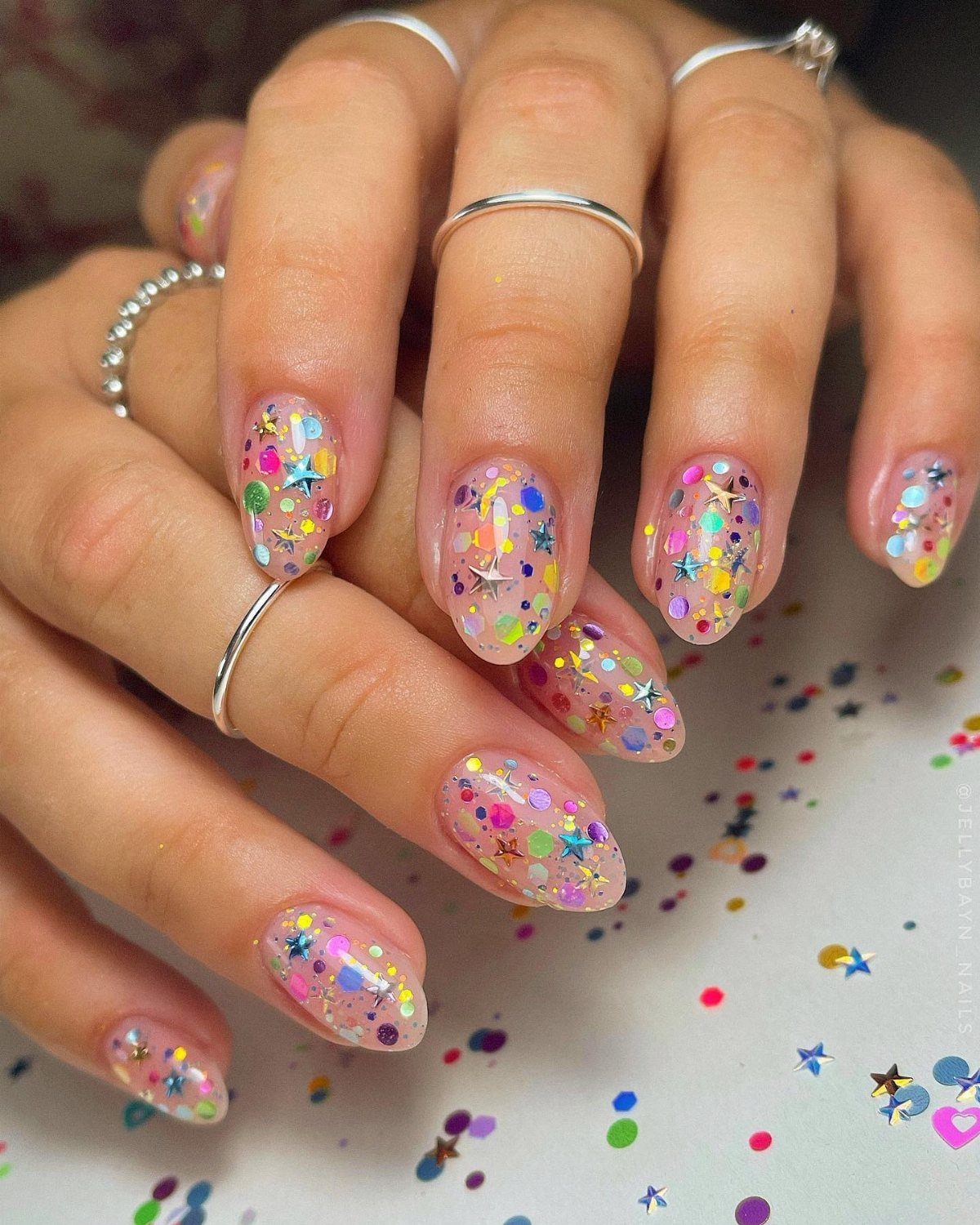 Hands with clear manicured nails decorated with colorful confetti glitter and silver rings.