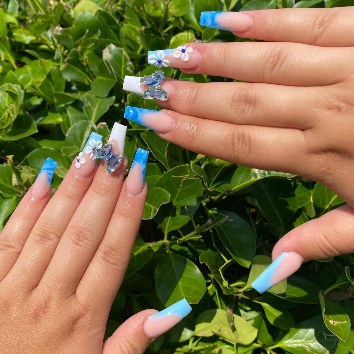 Long manicured nails with blue and white ombre, butterfly charms, and small white flowers.
