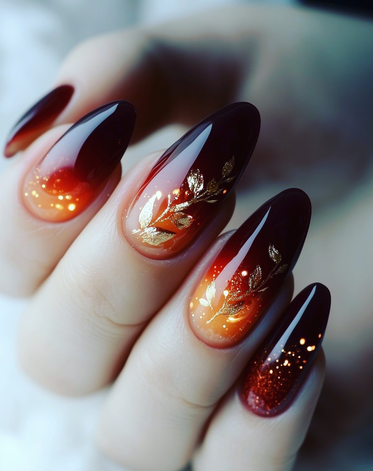 Long almond nails with dark burgundy and orange ombre and gold leaf accents.