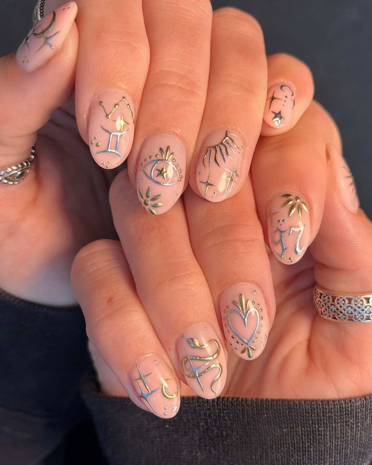 Hands with nude almond nails decorated with silver and gold celestial and zodiac symbols.