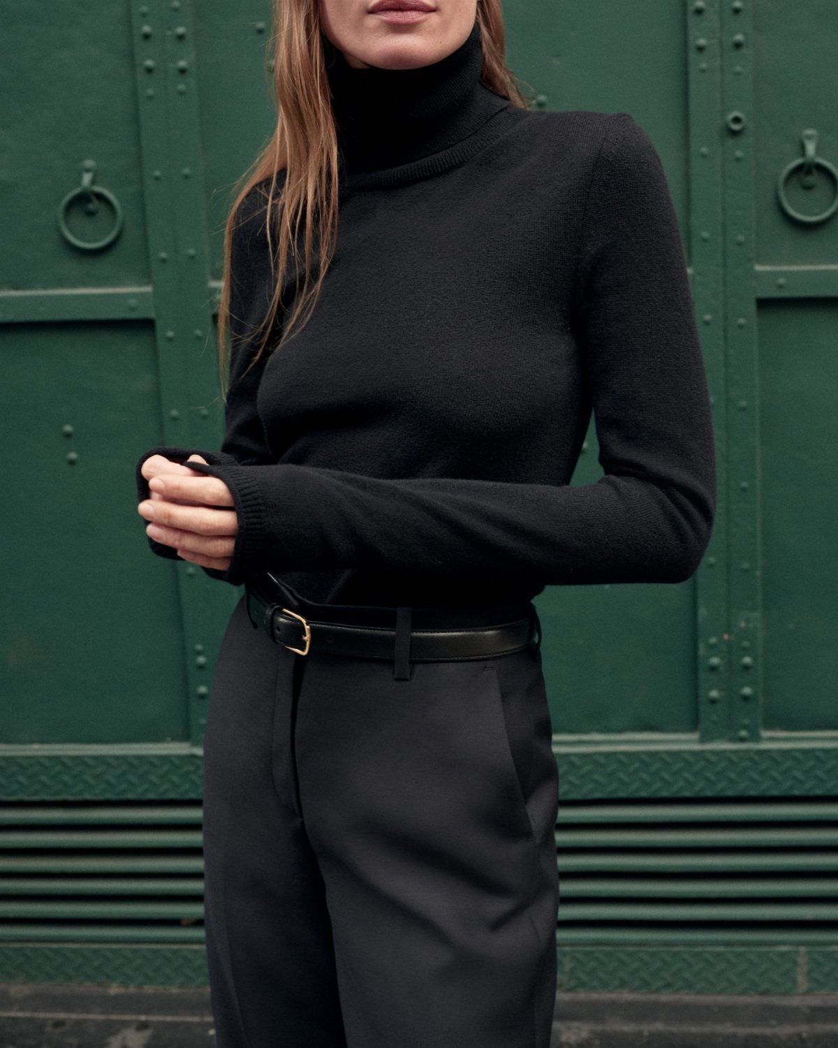 Woman wearing a black cashmere turtleneck and black trousers with a leather belt.