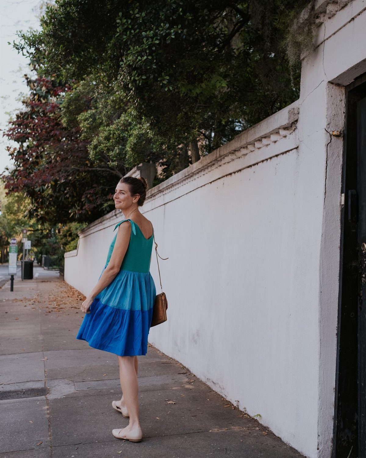 Woman in a bright blue and teal sundress walking along a sidewalk next to a white wall.