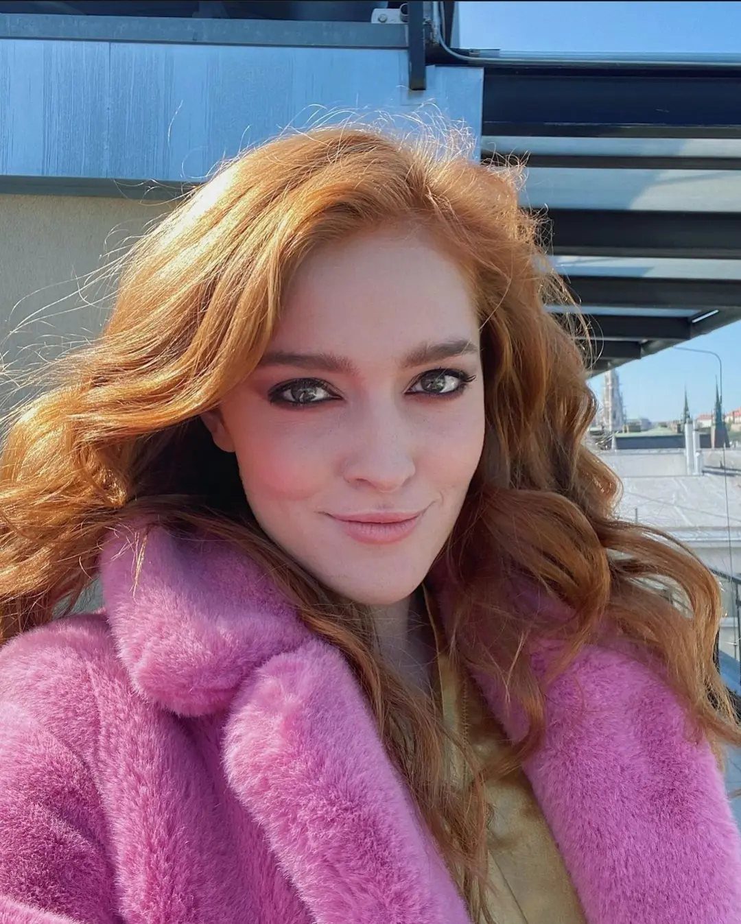 Red-haired woman smiling while wearing a voluminous pink faux fur coat outdoors.
