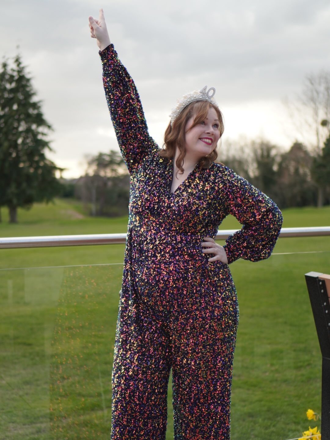 A woman in a colorful sequined jumpsuit and tiara posing on a balcony overlooking a park.