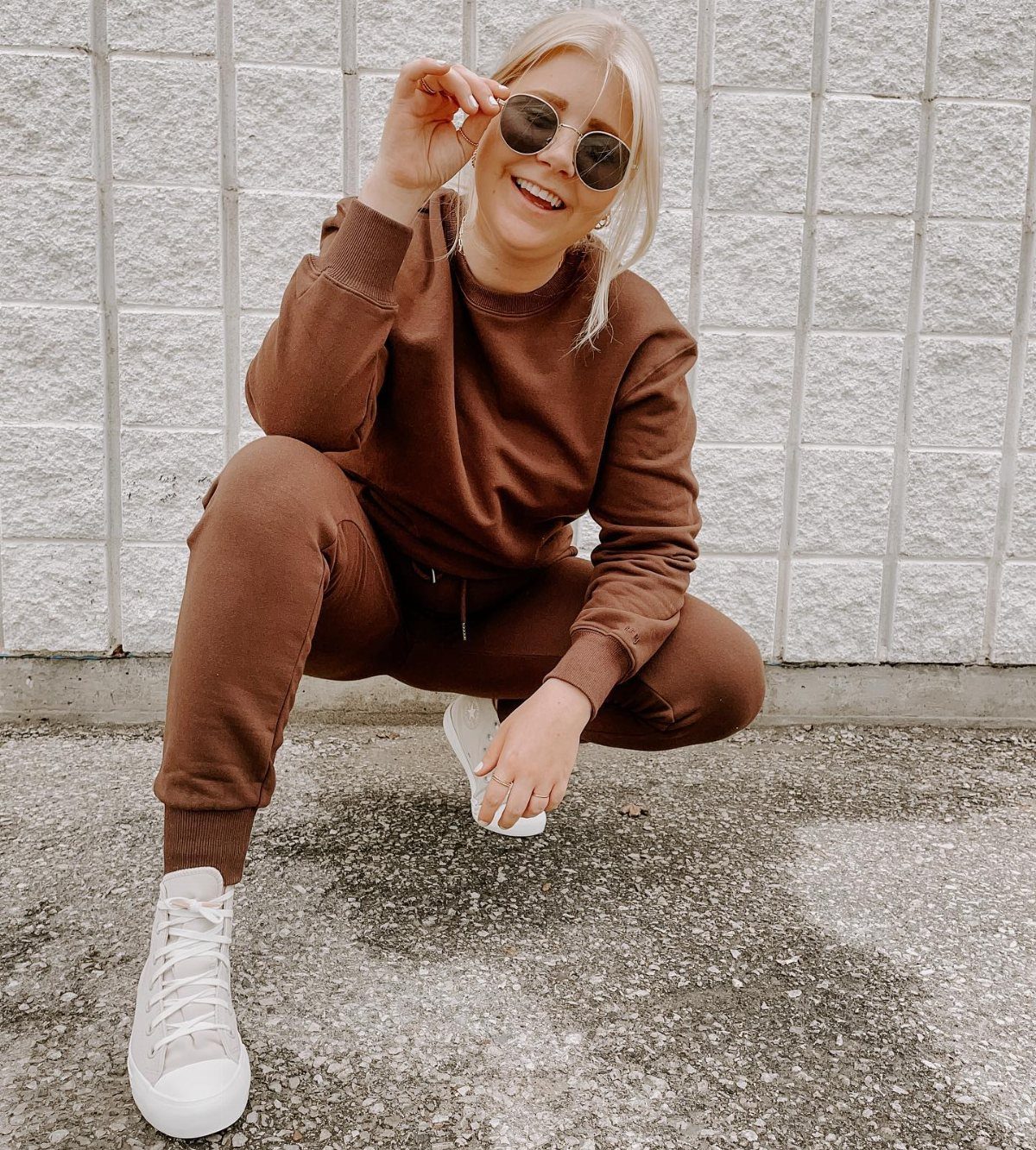 A blonde woman in a brown matching sweatsuit and white sneakers crouching outdoors.