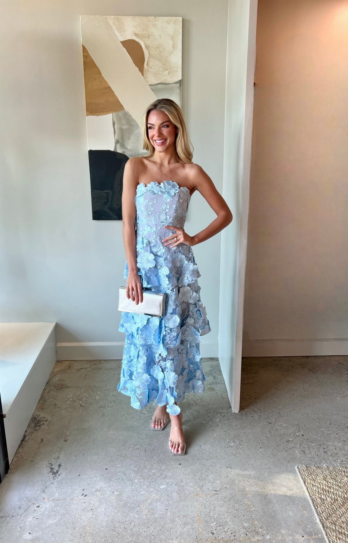 Smiling blonde woman posing in a light blue strapless floral midi dress and white clutch.