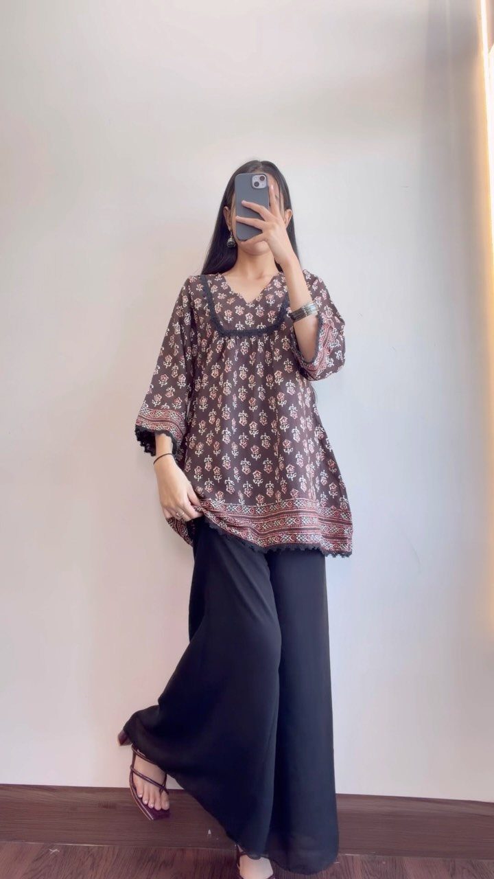 Woman wearing a patterned blouse and black wide-leg palazzo pants standing against a white wall.