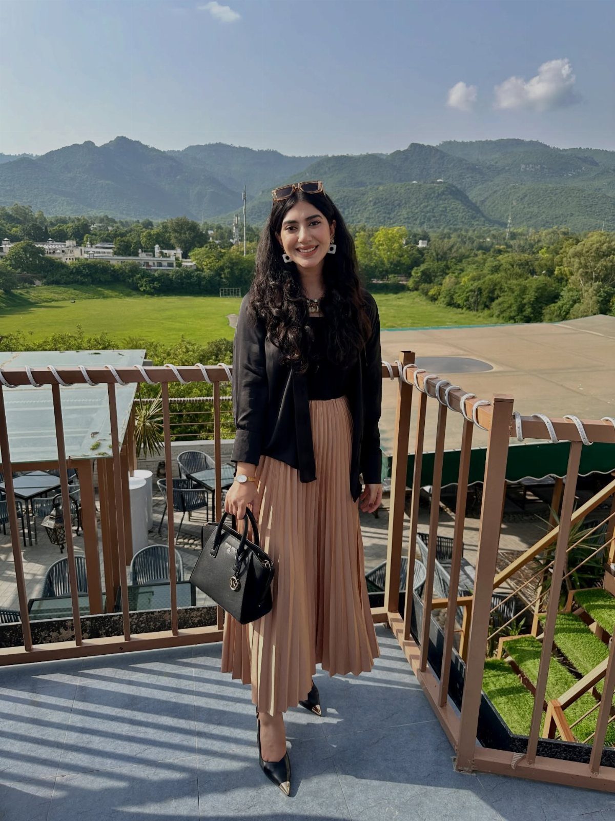 A woman wearing a black blouse and a tan pleated skirt standing on a balcony.