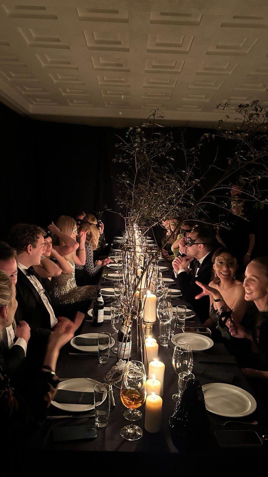 Guests in formal attire sit at a long, candlelit dinner table decorated with branches.