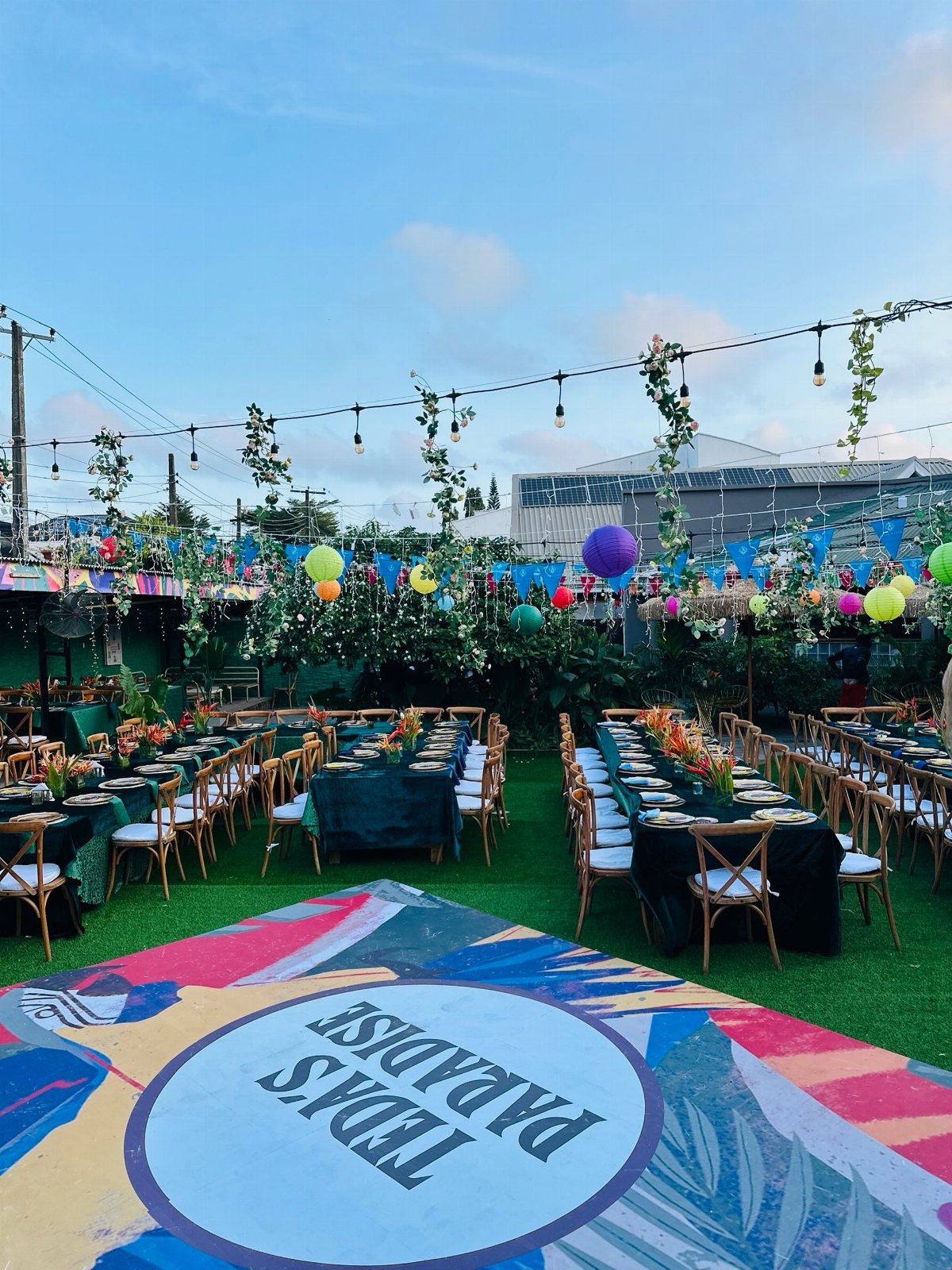 Long dining tables with green cloths and floral centerpieces set in an outdoor garden party space.
