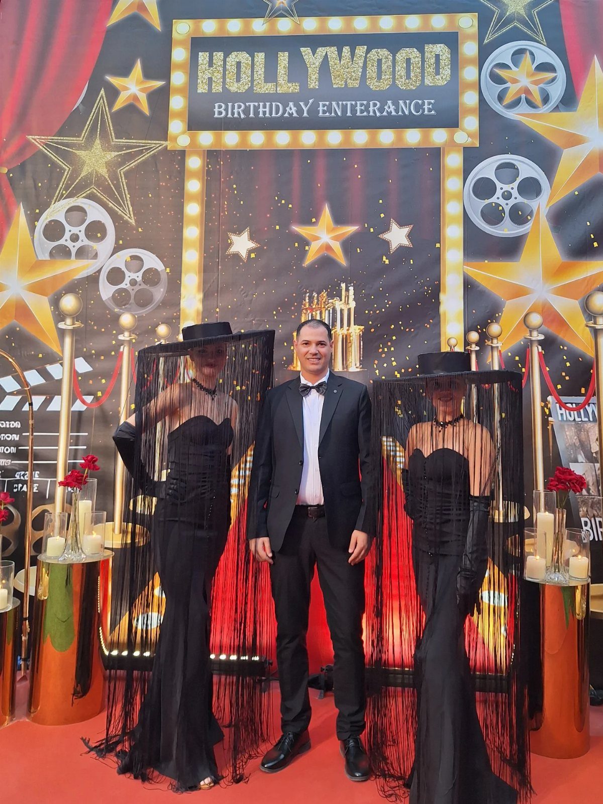 A man in a tuxedo standing between two women in black gowns and veils before a Hollywood backdrop.