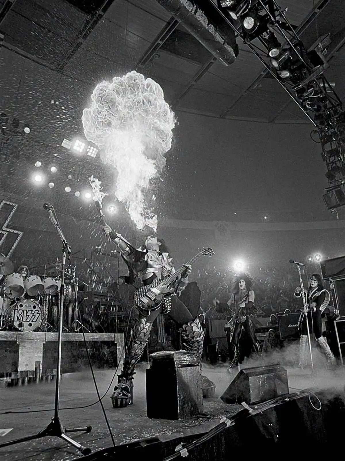 Rock band members performing on stage with large pyrotechnic fire effects.