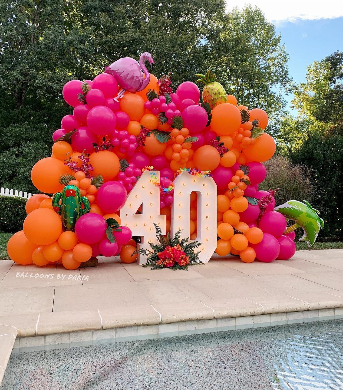 Large pink and orange balloon arch with tropical decorations surrounding white numbers forty by a pool.