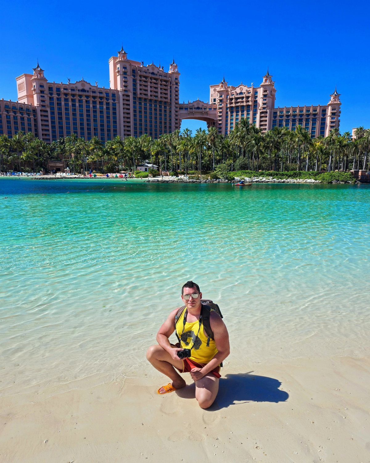 A man kneeling on a sandy beach with turquoise water and a large resort in the background.