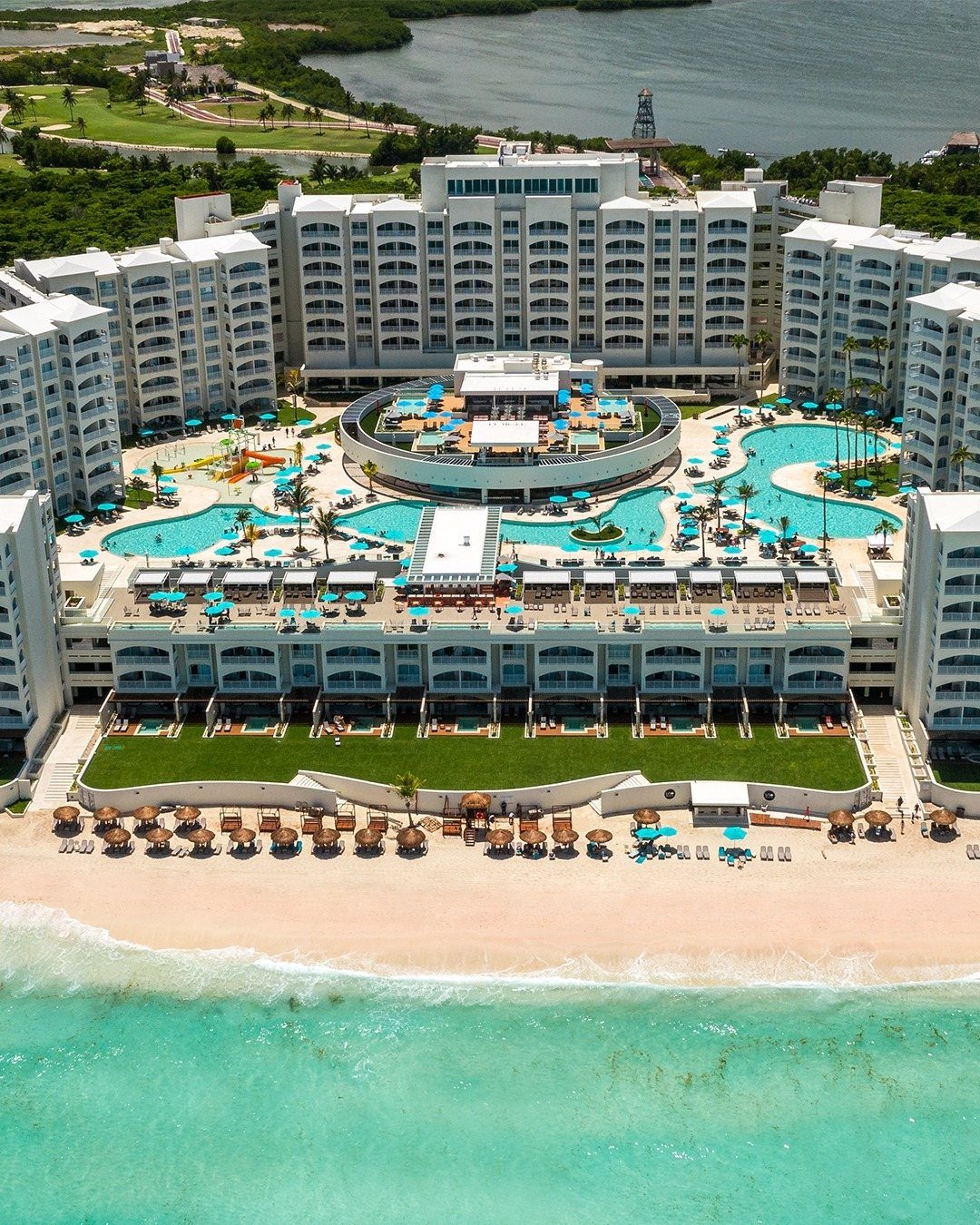 Aerial view of a large white luxury resort with turquoise pools and a sandy beach.