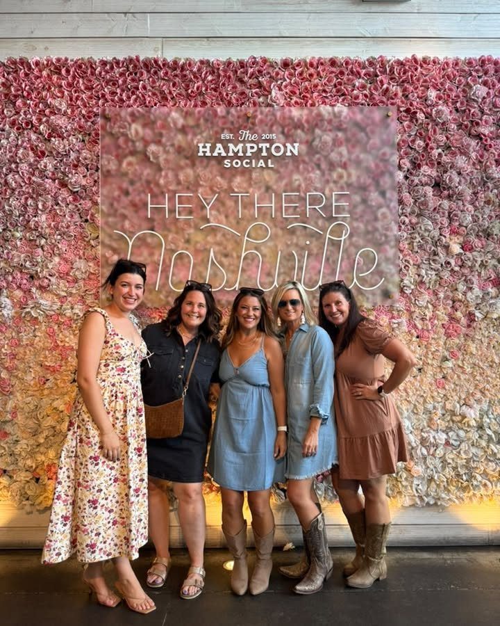 Five women smiling together in front of a pink rose wall with a Nashville sign.
