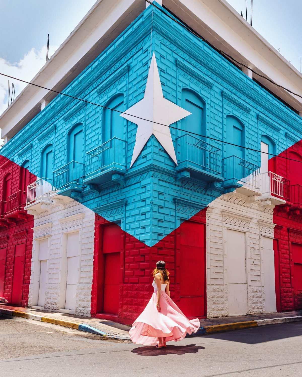 Woman in a pink dress standing before a building painted like the Puerto Rican flag.