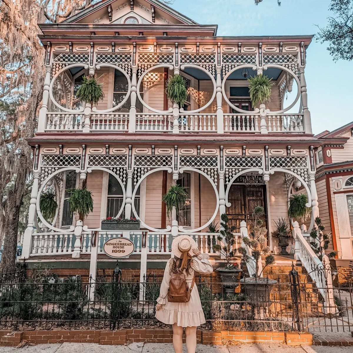 A woman stands before a historic two-story Victorian house with intricate white latticework and hanging plants.