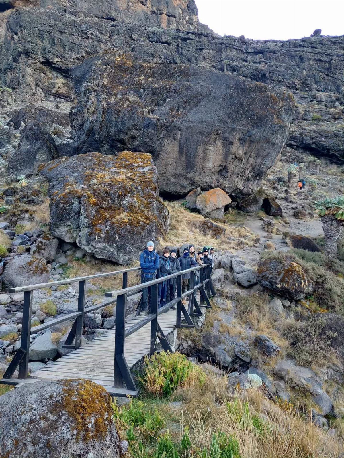 A group of hikers standing on a wooden bridge amidst large rocks and mountains.