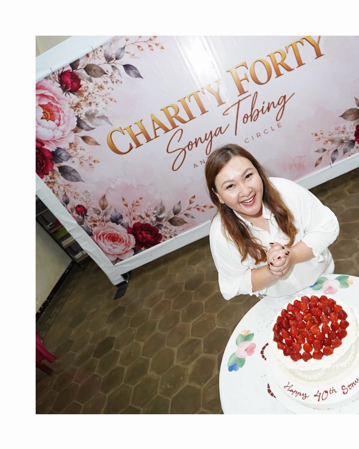 Smiling woman sitting behind a strawberry cake in front of a charity themed backdrop.