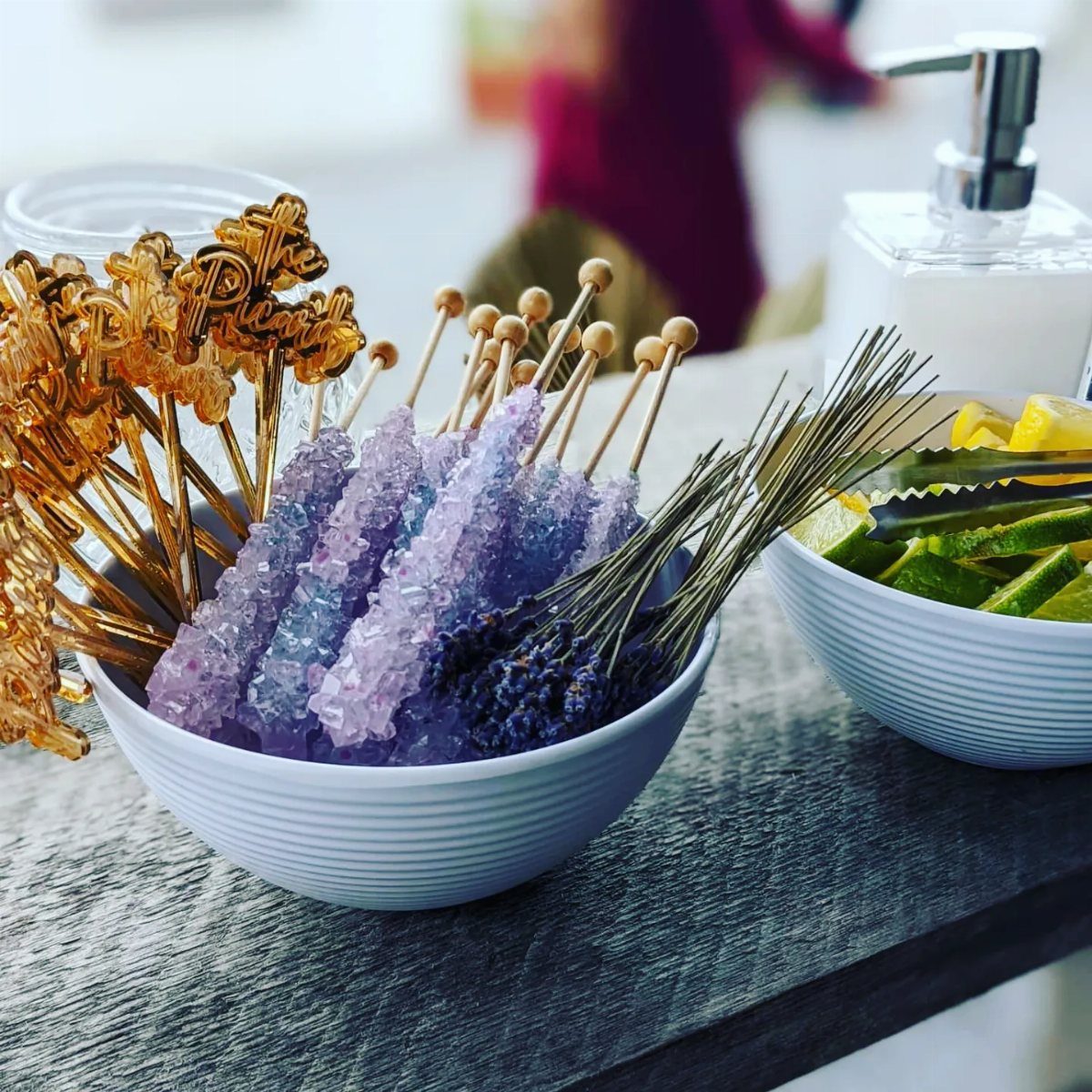 Purple sugar crystals, lavender, and gold cocktail picks arranged in white bowls on a wooden table.