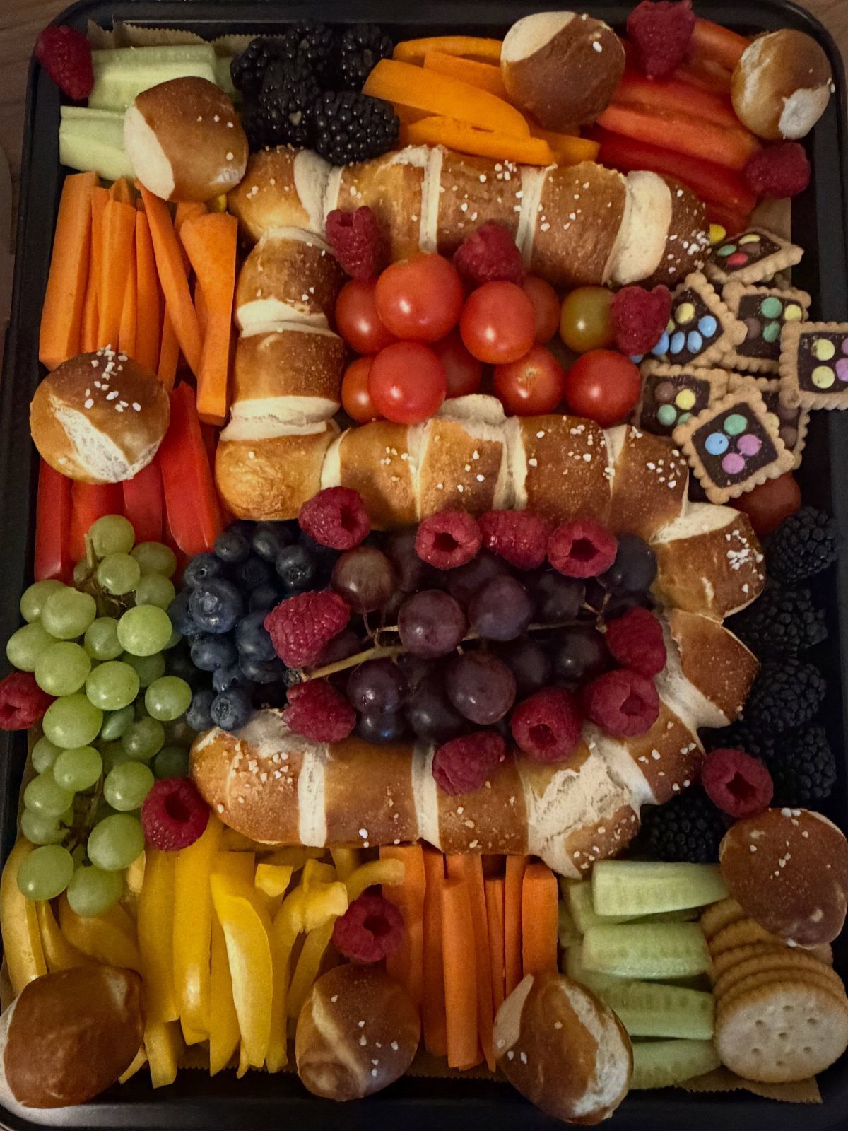 A large black tray overflowing with colorful fruits, vegetables, bread rolls, and cookies.