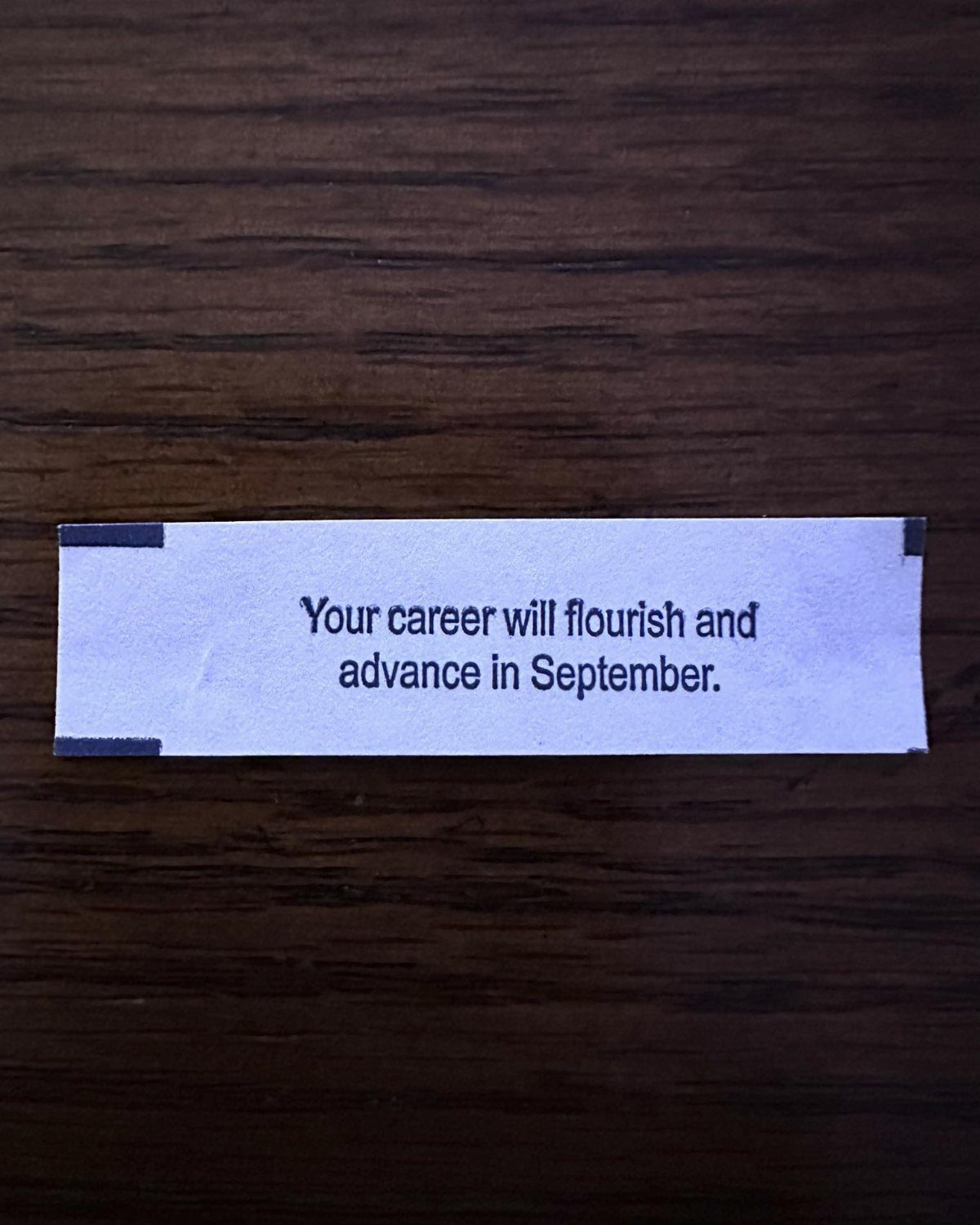 A white paper fortune cookie slip with blue text on a dark wood surface.
