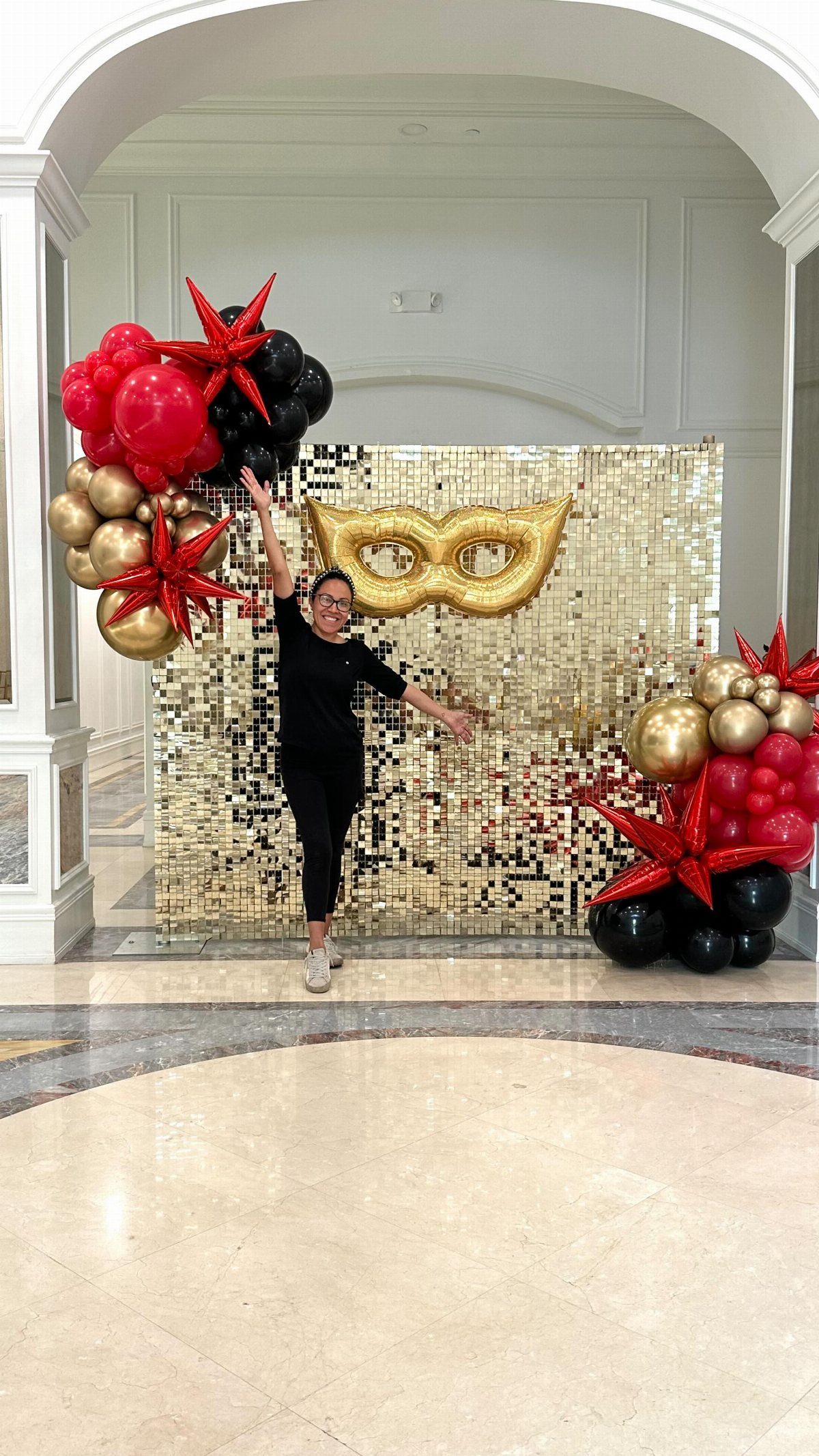 A woman posing in front of a gold sequin backdrop with a large gold mask and balloons.