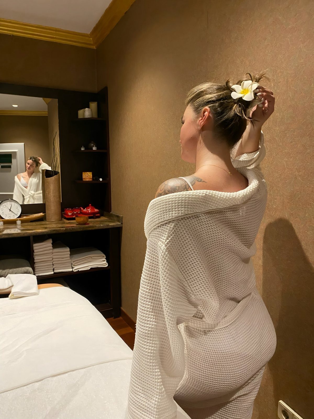 A woman in a white bathrobe with a flower in her hair stands in a spa room.