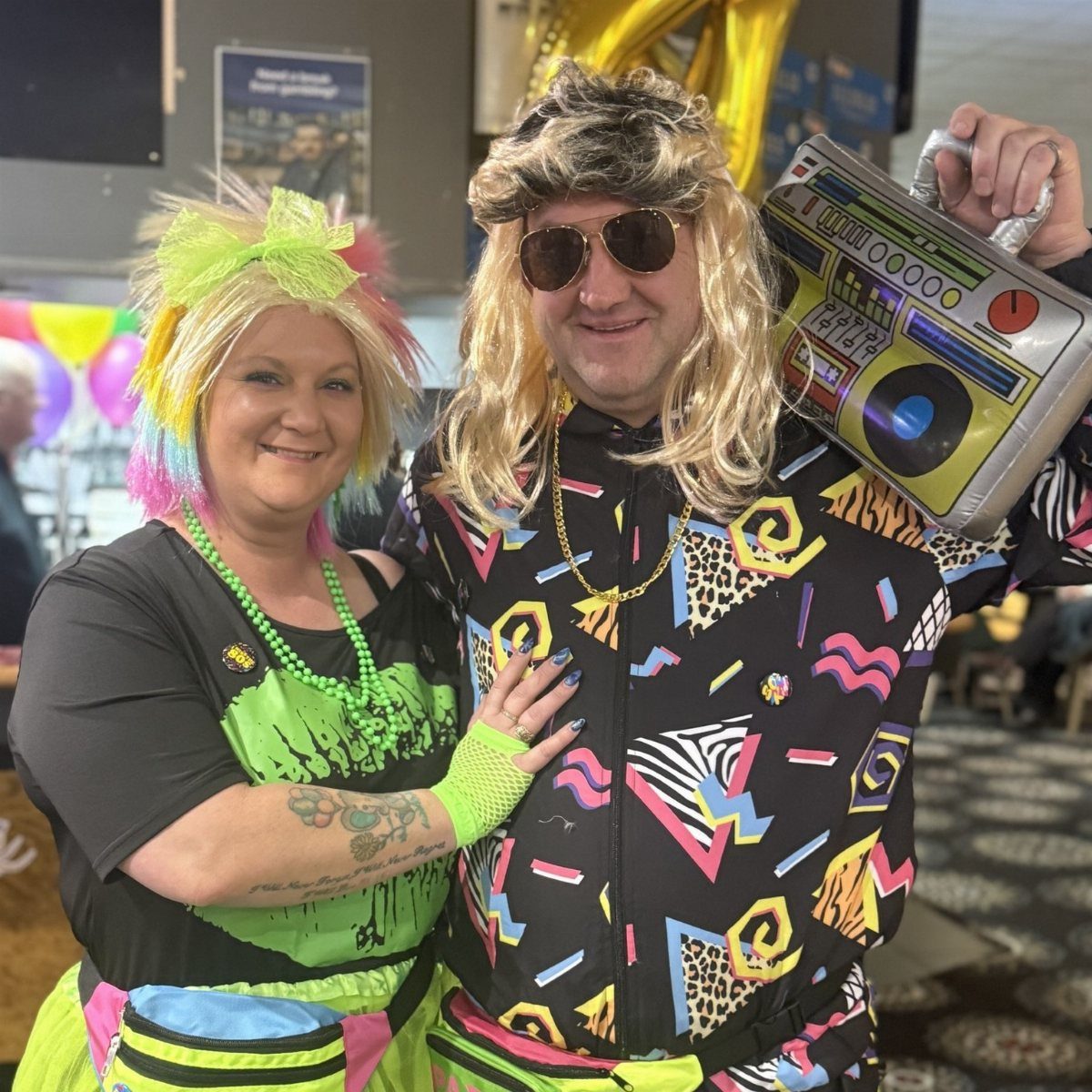 Two people smiling in bright, colorful 1980s-themed costumes holding a boombox prop.