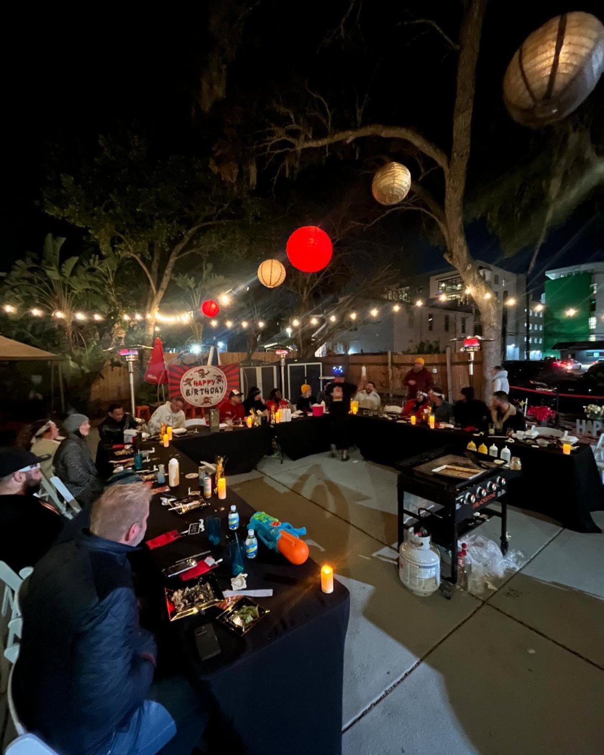People seated at long black tables decorated with candles and drinks under hanging paper lanterns.