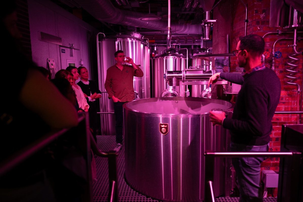 People observing a brewery tour amidst large stainless steel fermentation tanks and equipment.