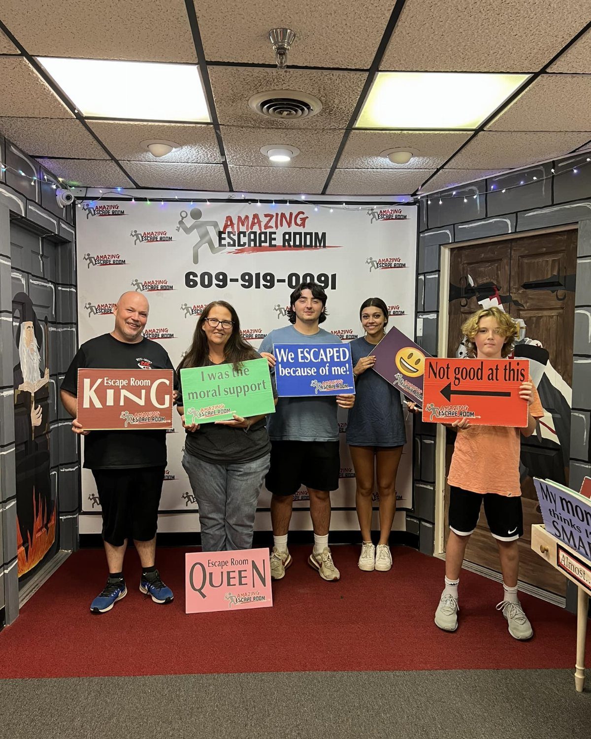 Five people posing with humorous signs in front of an Amazing Escape Room banner.
