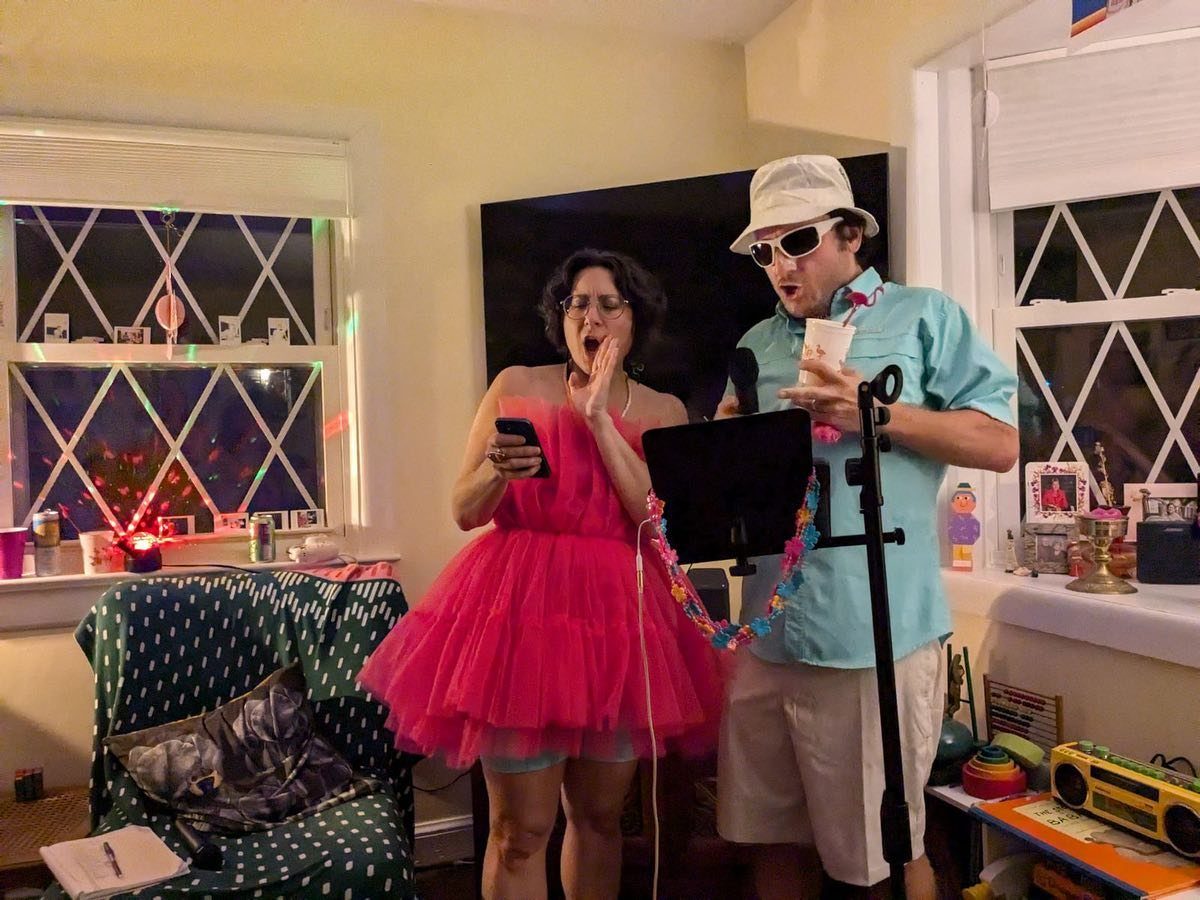 Two people in festive costumes singing and performing in a living room setting.