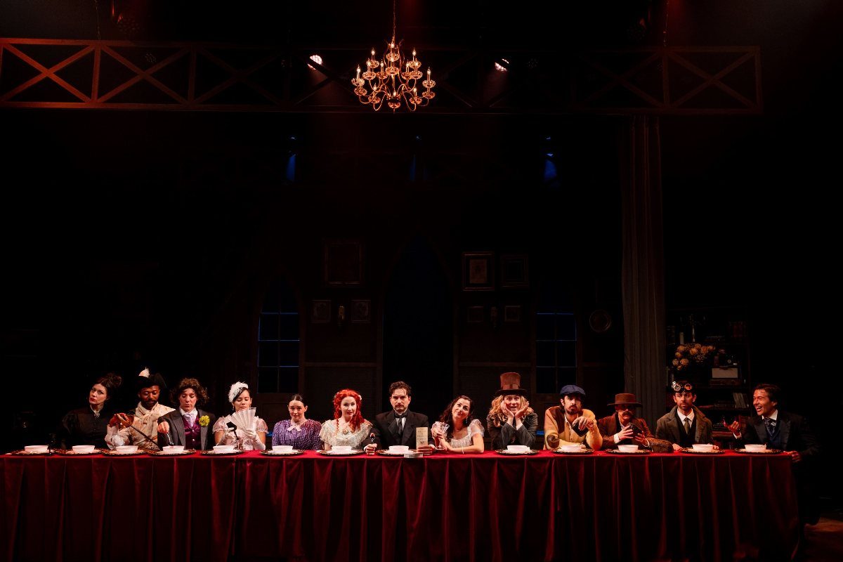People in vintage costumes sitting behind a long red table under a chandelier.
