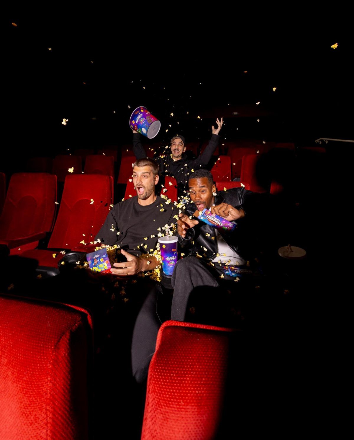 Three men celebrating in a movie theater with flying popcorn and snacks.