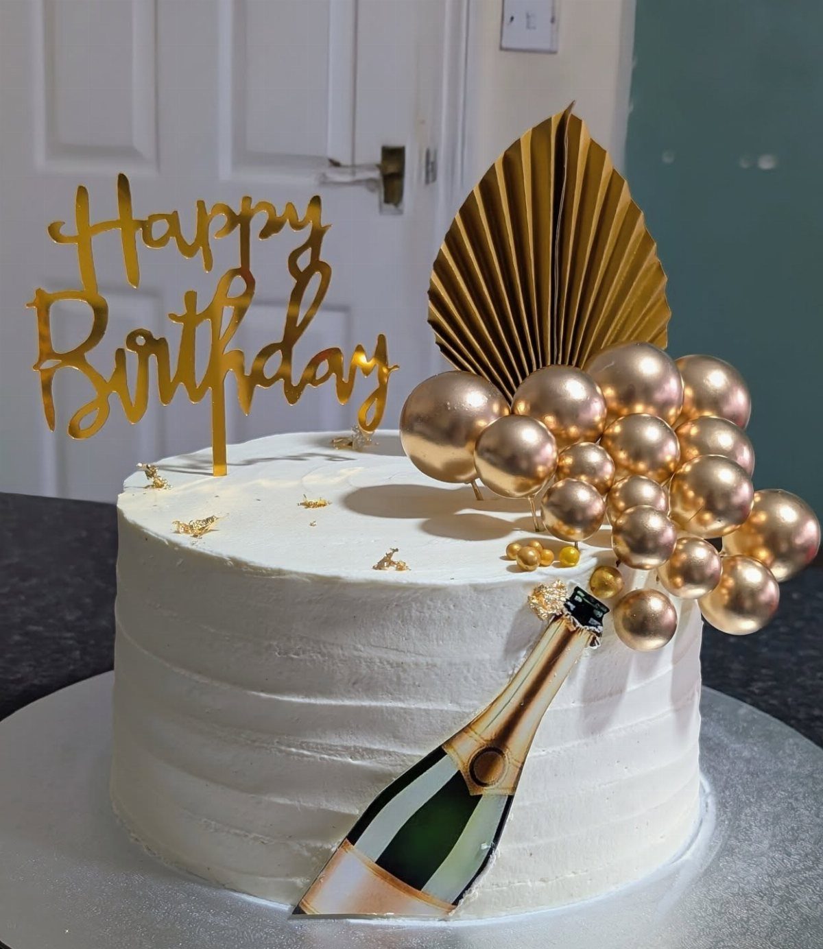 White buttercream cake decorated with gold birthday topper, champagne bottle cutout, and rose gold spheres.