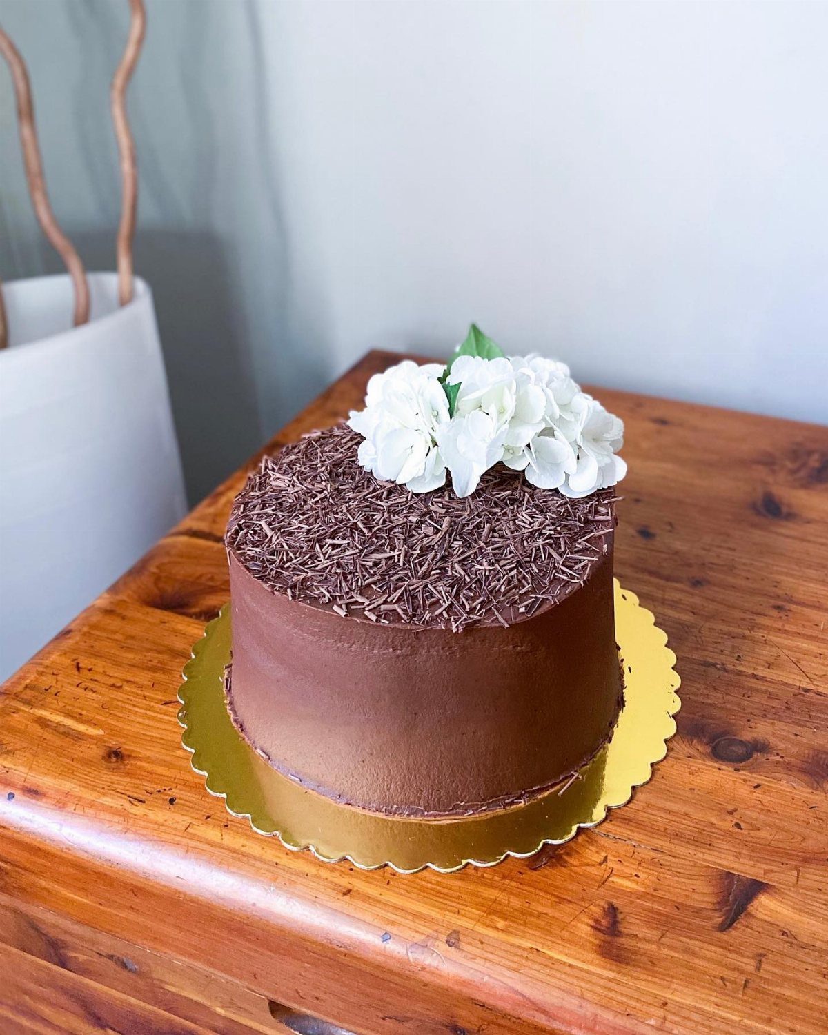 Chocolate cake topped with dark chocolate shavings and white hydrangea flowers on a wooden table.