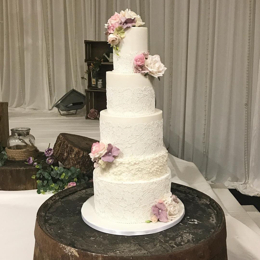 Four-tier white lace cake decorated with pink and white roses on a wooden stand.