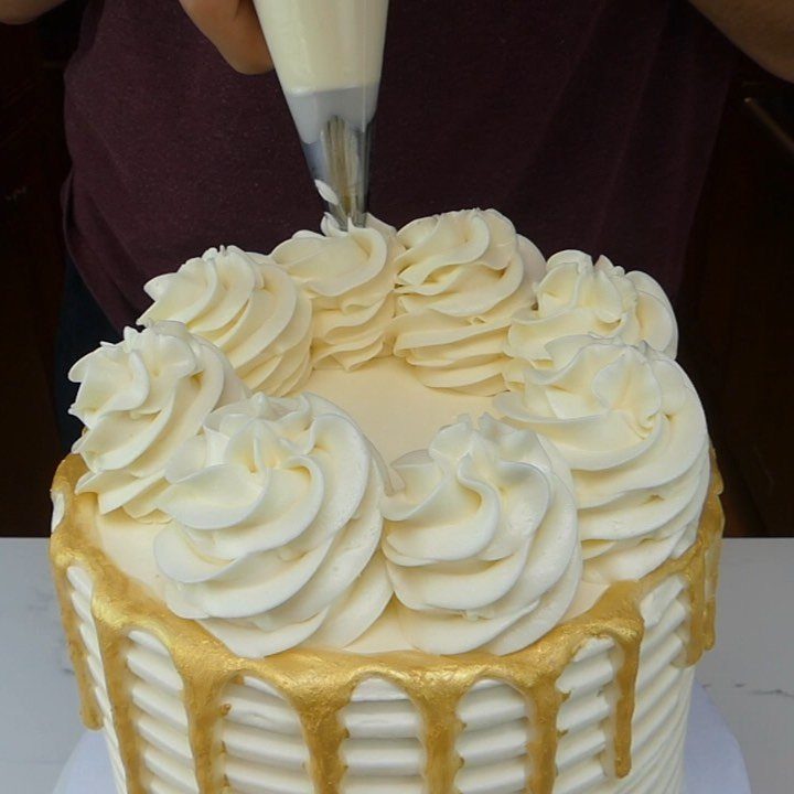 A white frosted cake with a metallic gold drip and large buttercream swirls on top.