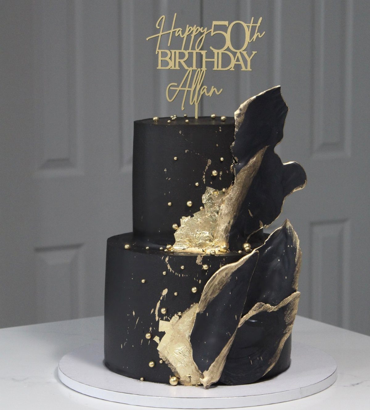 Two-tier matte black cake with gold leaf accents and a gold 50th birthday topper.