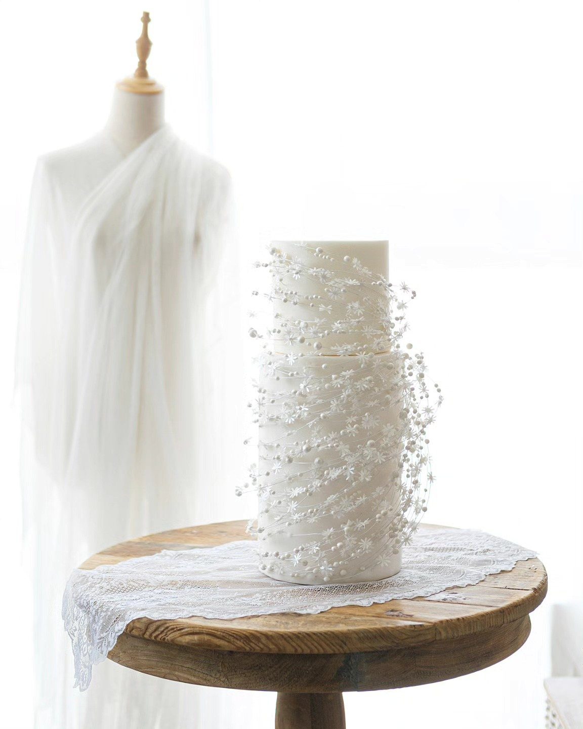 Two-tier white cake featuring intricate lace patterns and delicate edible pearls on a wooden table.