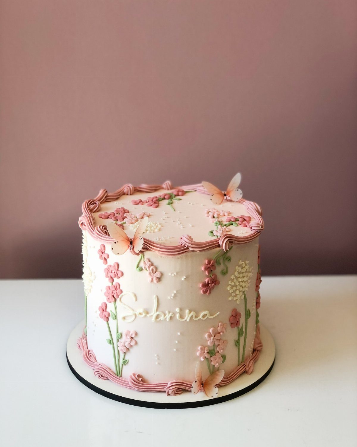 Pink buttercream cake decorated with delicate floral patterns, green stems, and small peach butterflies.