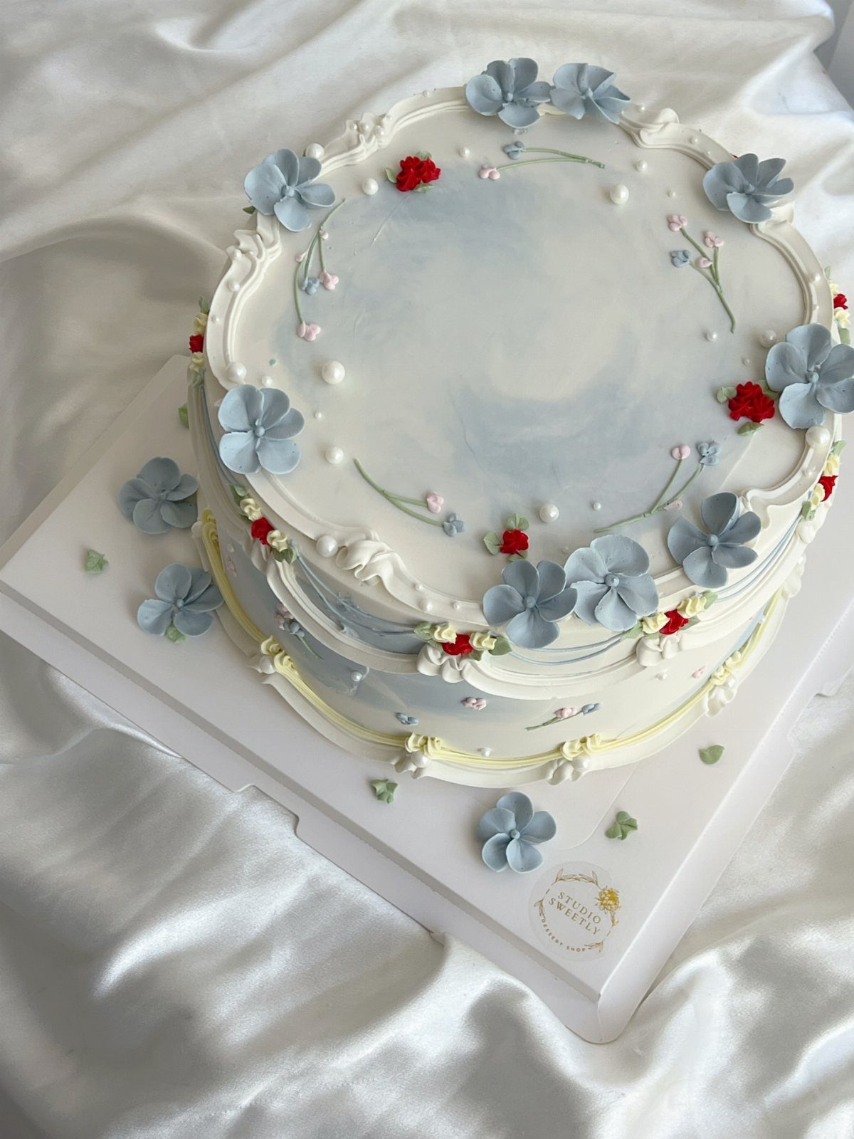 A light blue watercolor cake decorated with pale blue buttercream flowers and small red accents.
