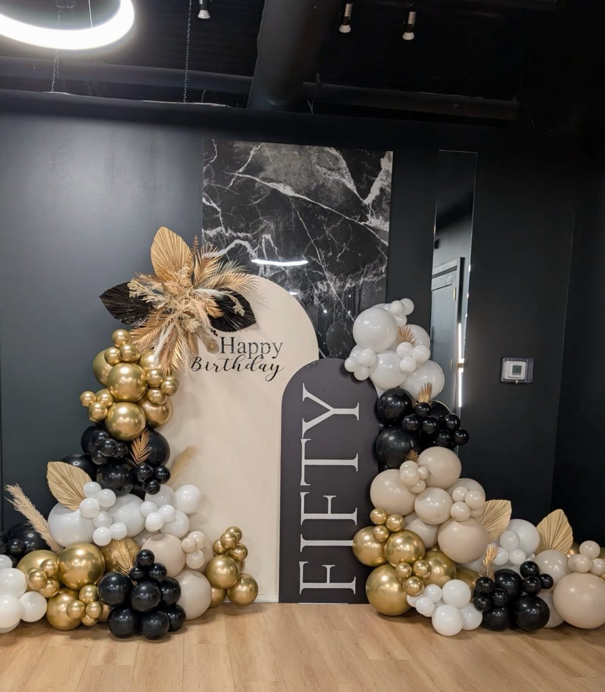 Black, gold, and white balloon arch surrounding birthday backdrops with Fifty and Happy Birthday text.
