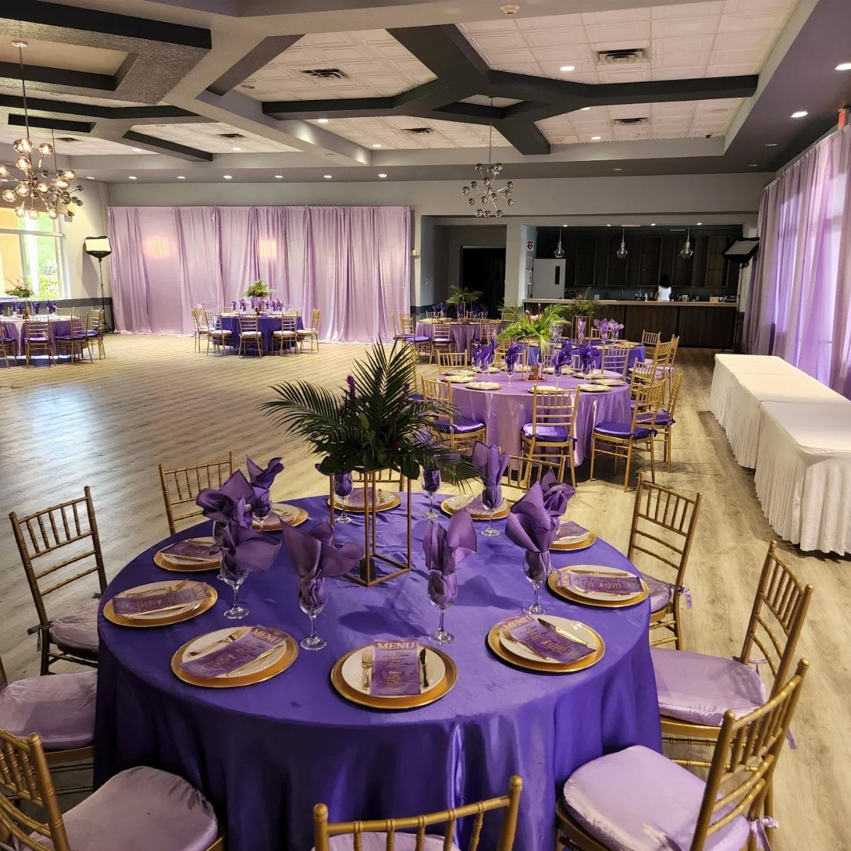 Purple banquet tables with gold plates and green palm leaf centerpieces in a large hall.